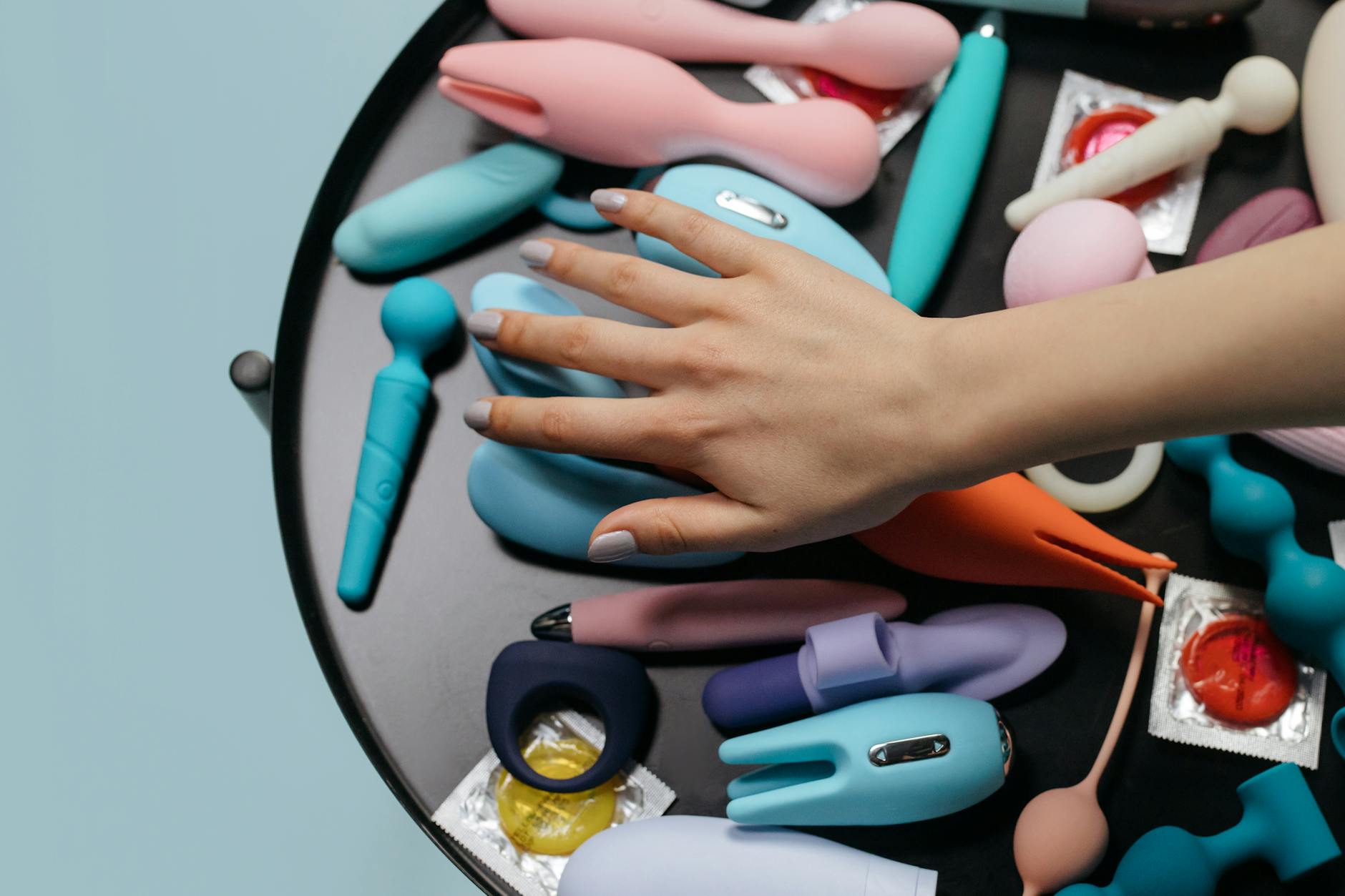 A hand reaching over a variety of colorful sex toys arranged on a table, showcasing different intimate options.